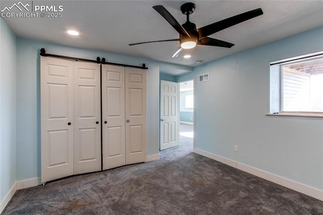 unfurnished bedroom featuring a barn door, ceiling fan, a closet, and dark colored carpet