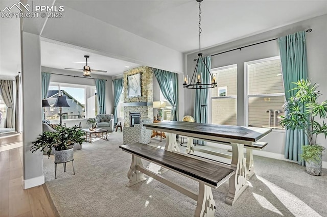 carpeted dining space with ceiling fan with notable chandelier, a stone fireplace, and a healthy amount of sunlight