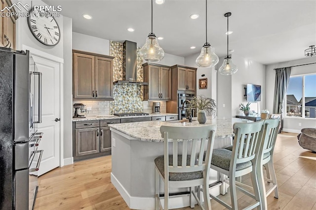 kitchen with appliances with stainless steel finishes, pendant lighting, an island with sink, light stone counters, and wall chimney exhaust hood