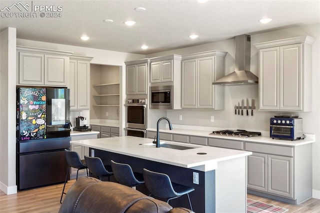 kitchen with sink, wall chimney range hood, light hardwood / wood-style flooring, an island with sink, and appliances with stainless steel finishes