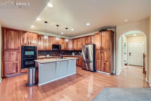 kitchen with an island with sink, hanging light fixtures, stainless steel fridge, light stone counters, and double oven