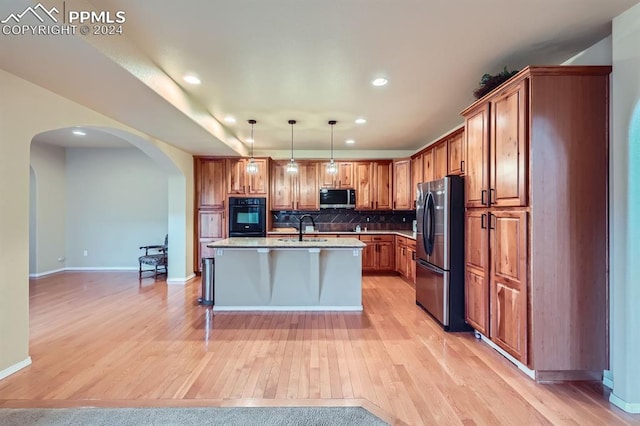 kitchen with light wood-type flooring, a kitchen island with sink, stainless steel appliances, hanging light fixtures, and backsplash