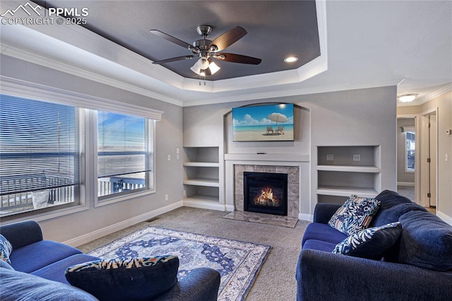 living room featuring a tile fireplace, a tray ceiling, carpet floors, ornamental molding, and built in shelves