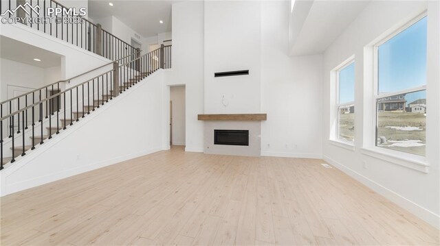 unfurnished living room with a towering ceiling, a glass covered fireplace, stairway, wood finished floors, and baseboards