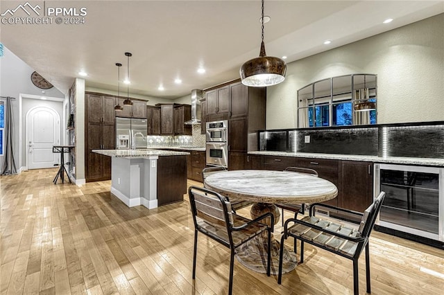 kitchen with light wood-type flooring, wall chimney exhaust hood, beverage cooler, a kitchen island with sink, and pendant lighting