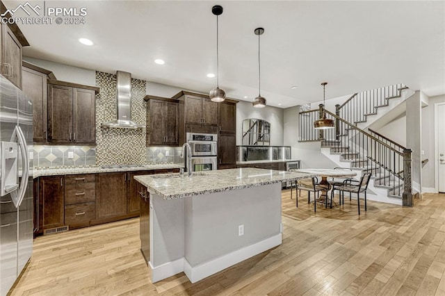 kitchen with stainless steel appliances, wall chimney range hood, decorative light fixtures, light hardwood / wood-style floors, and an island with sink