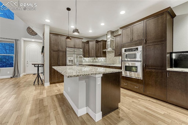 kitchen with appliances with stainless steel finishes, light stone counters, wall chimney exhaust hood, a breakfast bar, and a kitchen island with sink