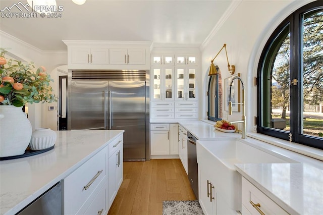 kitchen with appliances with stainless steel finishes, light stone counters, ornamental molding, light hardwood / wood-style floors, and white cabinetry