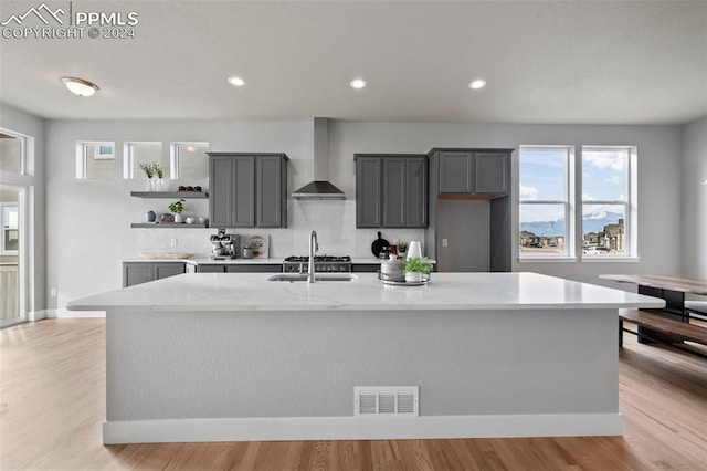 kitchen featuring a center island with sink, gray cabinets, wall chimney exhaust hood, and light hardwood / wood-style floors