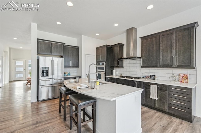 kitchen featuring a kitchen breakfast bar, light hardwood / wood-style flooring, wall chimney exhaust hood, an island with sink, and stainless steel appliances