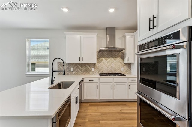 kitchen with sink, wall chimney exhaust hood, light wood-type flooring, white cabinetry, and stainless steel appliances