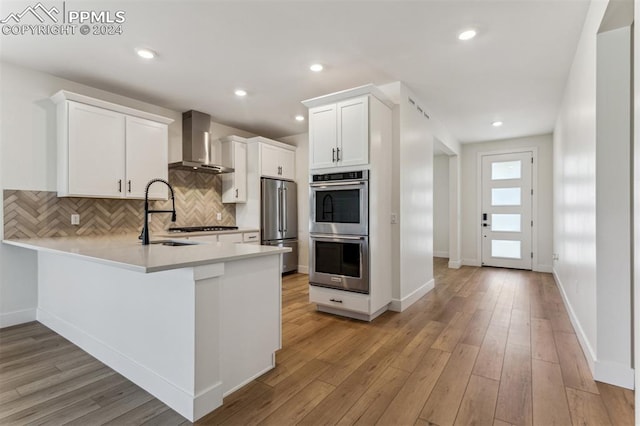 kitchen with kitchen peninsula, light wood-type flooring, wall chimney exhaust hood, stainless steel appliances, and white cabinets