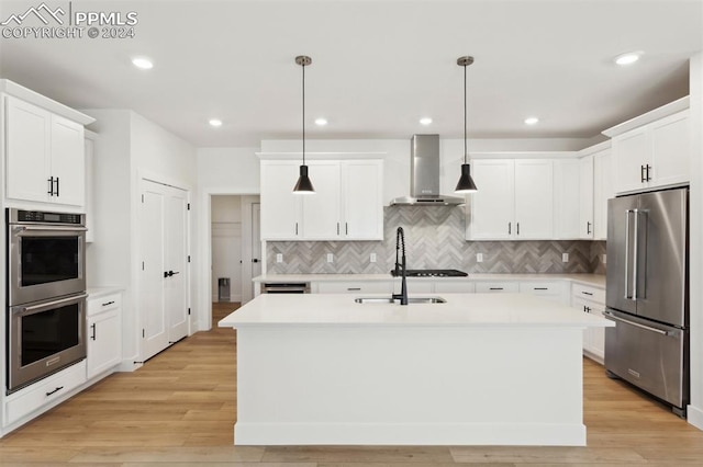 kitchen featuring decorative light fixtures, wall chimney range hood, stainless steel appliances, and an island with sink