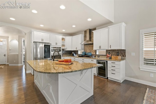 kitchen featuring wine cooler, wall chimney exhaust hood, an island with sink, appliances with stainless steel finishes, and white cabinetry