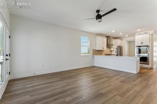 kitchen with kitchen peninsula, appliances with stainless steel finishes, wall chimney range hood, light hardwood / wood-style floors, and white cabinetry