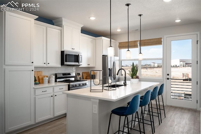 kitchen featuring stainless steel appliances, white cabinets, hardwood / wood-style floors, hanging light fixtures, and an island with sink