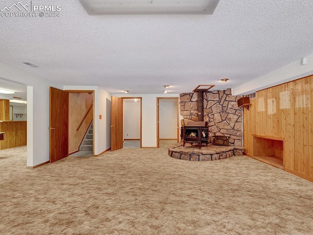 unfurnished living room featuring a wood stove, wooden walls, light carpet, and a textured ceiling