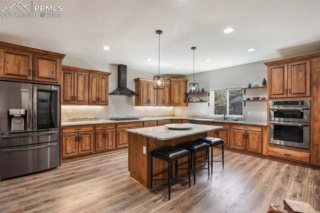 kitchen featuring a center island, wall chimney range hood, hanging light fixtures, light wood-type flooring, and appliances with stainless steel finishes