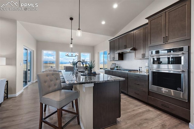 kitchen with stainless steel appliances, dark stone countertops, decorative light fixtures, a kitchen bar, and a kitchen island with sink