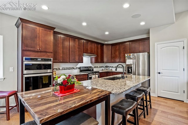 kitchen featuring a kitchen island with sink, sink, light wood-type flooring, light stone counters, and stainless steel appliances