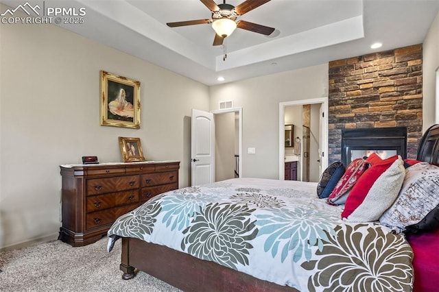 carpeted bedroom featuring ceiling fan, a fireplace, a tray ceiling, and ensuite bath