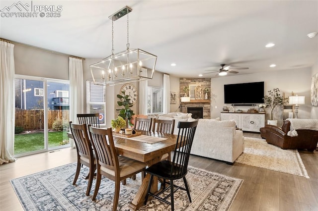 dining area with ceiling fan with notable chandelier, hardwood / wood-style flooring, a stone fireplace, and a wealth of natural light