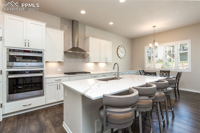 kitchen with sink, an island with sink, white cabinetry, and wall chimney range hood