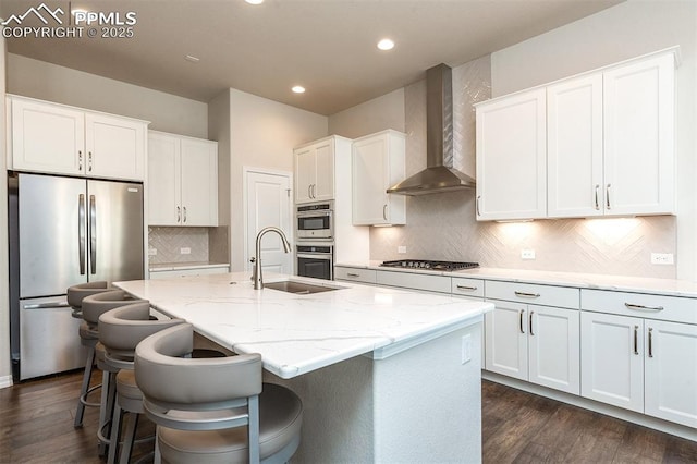 kitchen featuring white cabinets, a kitchen island with sink, wall chimney exhaust hood, and appliances with stainless steel finishes