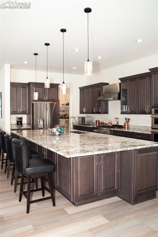 kitchen with wall chimney exhaust hood, dark brown cabinetry, stainless steel appliances, a large island with sink, and decorative light fixtures