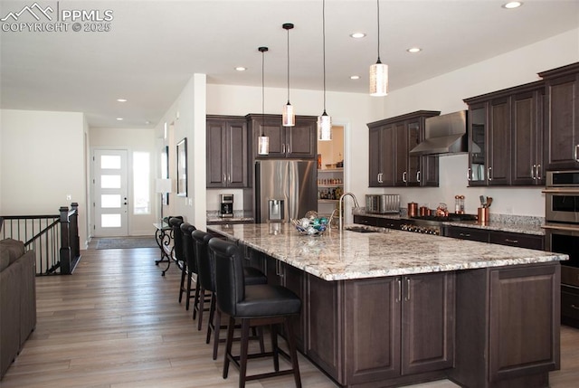 kitchen featuring stainless steel appliances, a spacious island, sink, wall chimney range hood, and hanging light fixtures