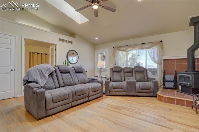 living room with a wood stove, ceiling fan, lofted ceiling with skylight, and light wood-type flooring