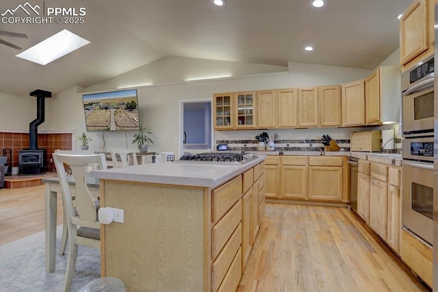 kitchen with stainless steel appliances, light brown cabinetry, light wood finished floors, lofted ceiling with skylight, and a wood stove