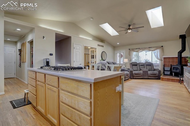 kitchen with vaulted ceiling with skylight, light wood-style flooring, light brown cabinetry, a wood stove, and stainless steel gas stovetop