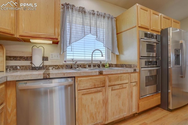 kitchen with tile counters, stainless steel appliances, light brown cabinetry, light wood-style floors, and a sink