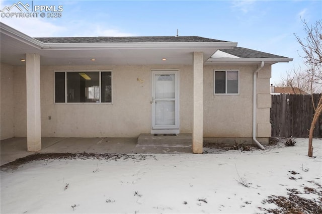 snow covered property entrance featuring roof with shingles, stucco siding, fence, and a patio