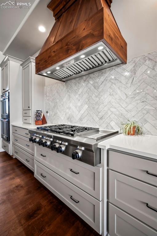 kitchen featuring tasteful backsplash, multiple ovens, custom range hood, stainless steel gas cooktop, and dark hardwood / wood-style floors