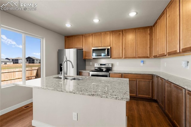 kitchen with a kitchen island with sink, dark wood-type flooring, sink, light stone countertops, and appliances with stainless steel finishes