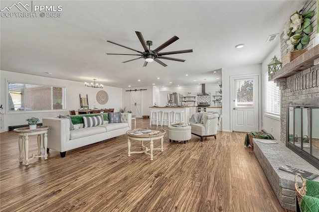 living room featuring a barn door, a large fireplace, wood-type flooring, and ceiling fan with notable chandelier