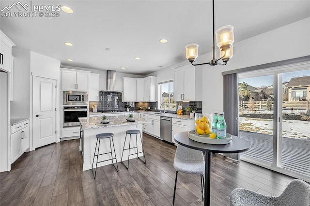 kitchen featuring pendant lighting, wall chimney range hood, appliances with stainless steel finishes, white cabinetry, and light stone counters