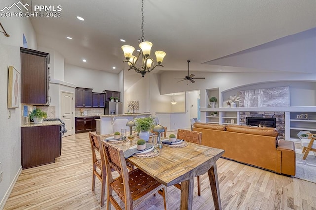 dining area with built in shelves, a stone fireplace, high vaulted ceiling, ceiling fan with notable chandelier, and light wood-type flooring