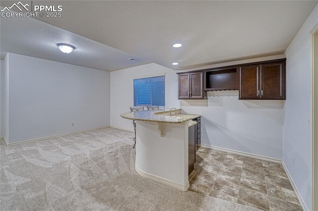 kitchen featuring dark brown cabinetry, light stone countertops, kitchen peninsula, light colored carpet, and a breakfast bar area