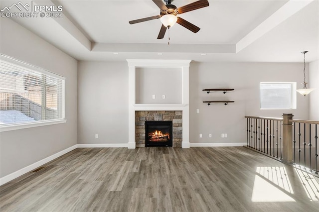 unfurnished living room featuring ceiling fan, a stone fireplace, wood-type flooring, and a tray ceiling