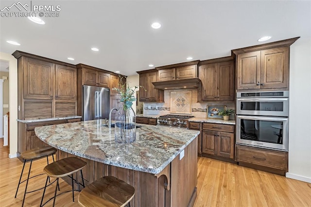 kitchen with sink, stainless steel appliances, light stone counters, light hardwood / wood-style floors, and a center island with sink