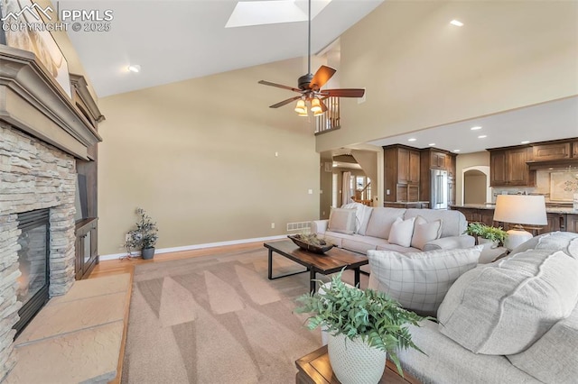 living room with a stone fireplace, ceiling fan, high vaulted ceiling, and a skylight