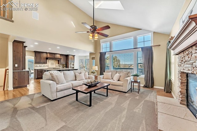 living room with a skylight, ceiling fan, a stone fireplace, high vaulted ceiling, and light hardwood / wood-style floors