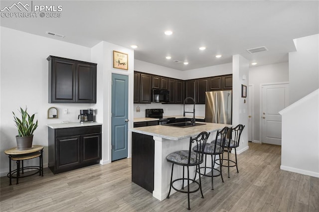 kitchen with a breakfast bar, a kitchen island with sink, black appliances, sink, and light wood-type flooring