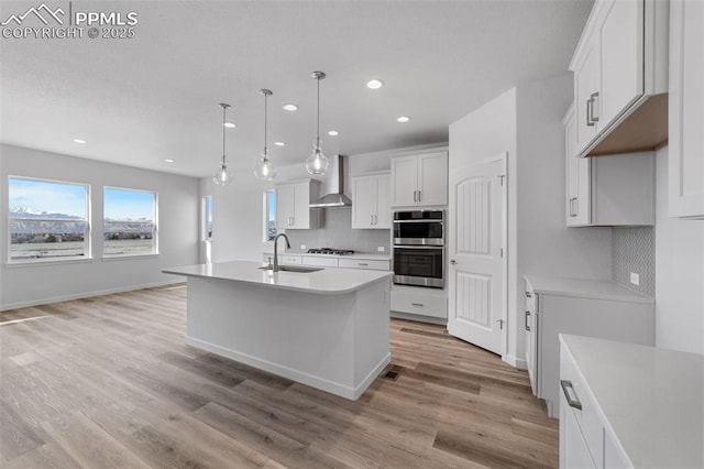 kitchen featuring a kitchen island with sink, wall chimney range hood, sink, decorative light fixtures, and white cabinetry