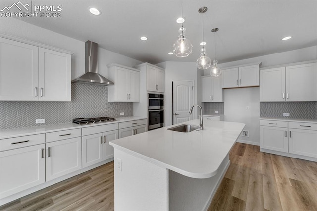 kitchen with appliances with stainless steel finishes, wall chimney exhaust hood, sink, a center island with sink, and white cabinetry