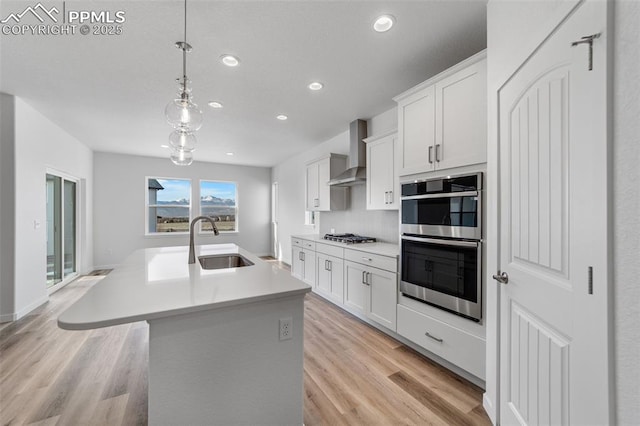 kitchen featuring sink, wall chimney exhaust hood, an island with sink, decorative light fixtures, and white cabinets