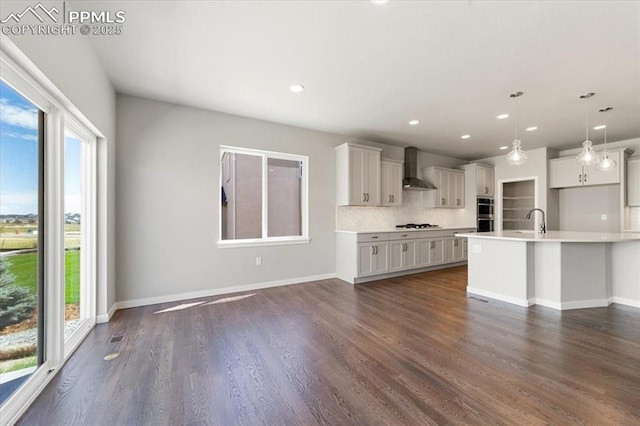 kitchen featuring wall chimney exhaust hood, a kitchen island with sink, dark wood-type flooring, pendant lighting, and decorative backsplash